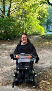 Catharina Wesemüller sitzt in ihrem E-Rolli. Sie ist schwarz gekleidet und im Hintergrund sind grüne Büsche.