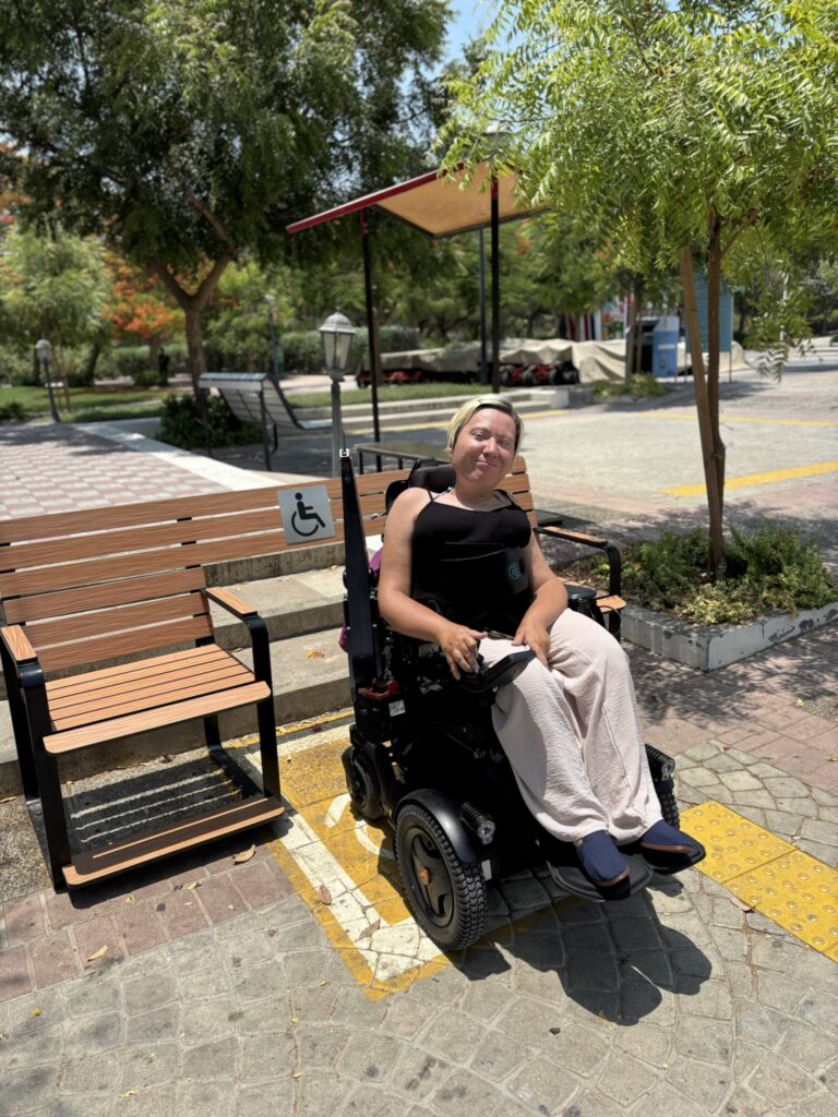 Foto von Jenny Bießmann. Jenny sitzt in einem Elektrorollstuhl in einer Lücke einer barrierefreien Parkbank - rechts und links von ihr sind Sitzplätze für Fußgänger.